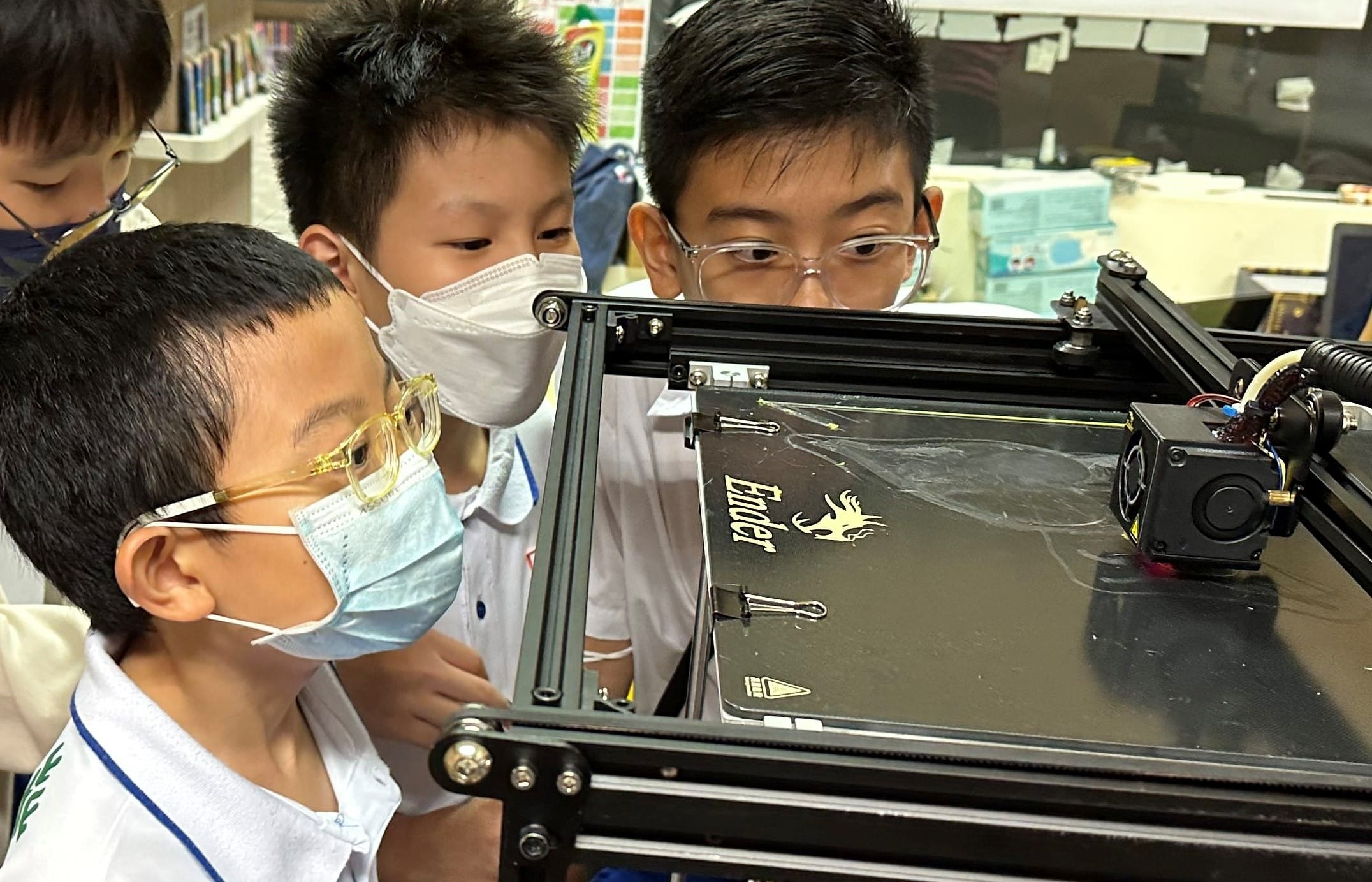 Three young students wearing face masks and glasses lean in closely to observe an Ender 3D printer in a classroom.
